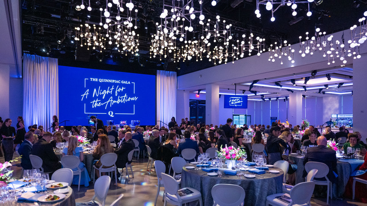 People sitting at dining tables in a large room with chandeliers and Gala signage on a screen