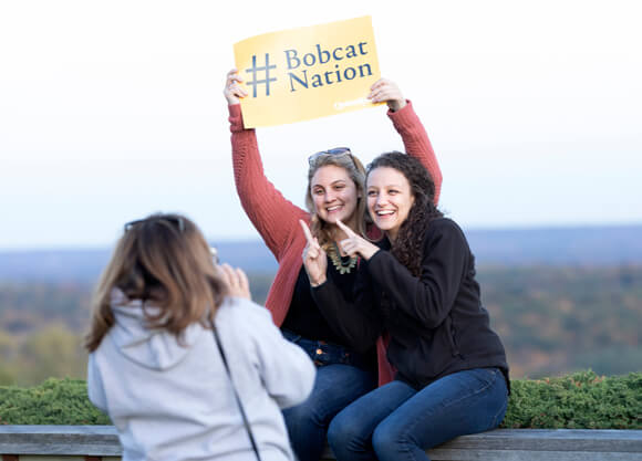Two alumni pose for a photo sitting on a wall at Rocky Top holding a #Bobcat Nation sign