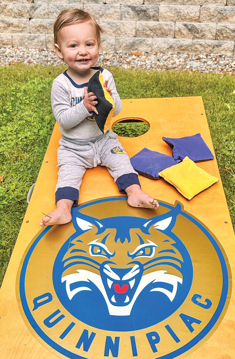 Baby Rigazio in a Bobcats outfit smiles and sits on a Quinnipiac bean bag toss game board