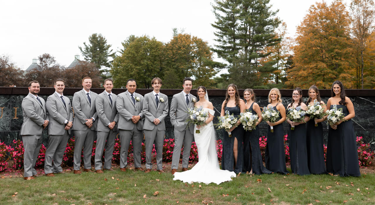 Julia Basso and Cameron Mowry smile with their wedding party in front of a Quinnipiac sign